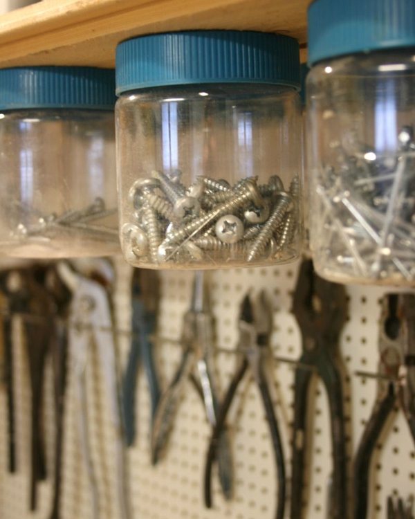 Clear jars with blue lids screwed to the underside of a workshop shelf, screws inside, pliers hanging on pegboard below